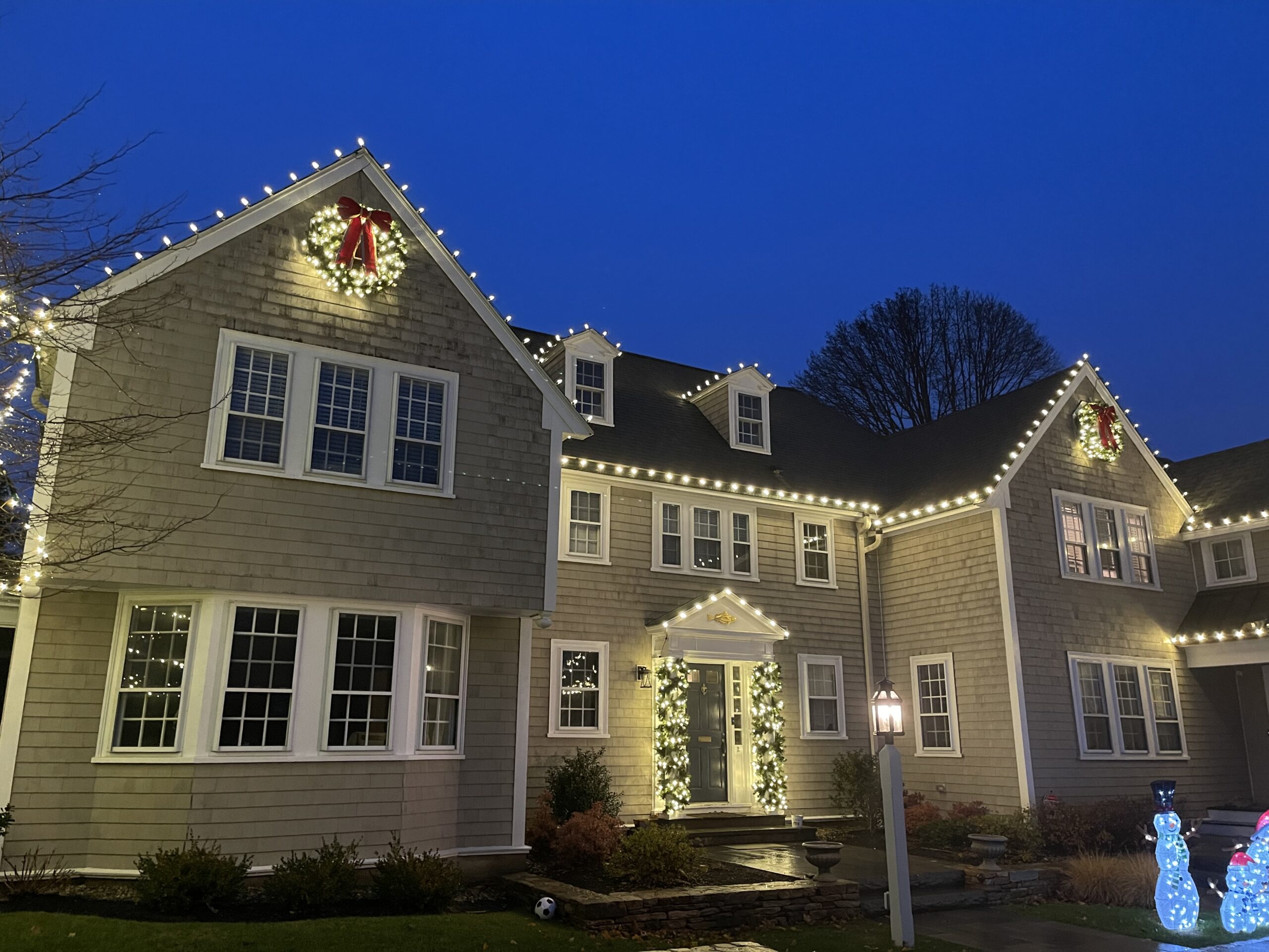A large two-story house decorated with white string lights along the roof and windows, two red wreath bows, and glowing snowman figures on the lawn, all illuminated at dusk.