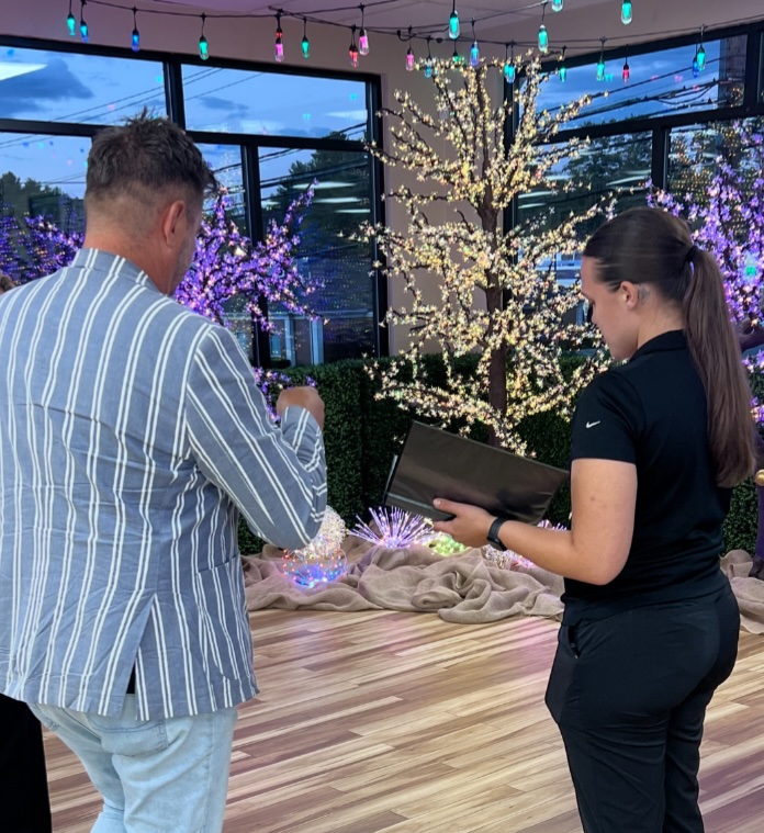 A man in a striped blazer and a woman in a black outfit stand indoors, admiring illuminated decorative trees and lights—perfect for highlighting wholesale benefits in décor settings, with the woman holding a black folder.