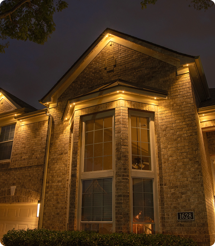 Permanent warm yellow roofline lighting two story brick house large windows NEI Distributors North Reading MA.